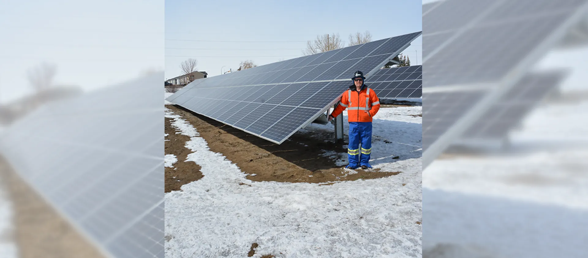 A SaskEnergy employee wearing an orange coat, blue pants and black hardhat, stands in front of a solar panel installation. Some snow is on the ground, with brown grass peeking through.