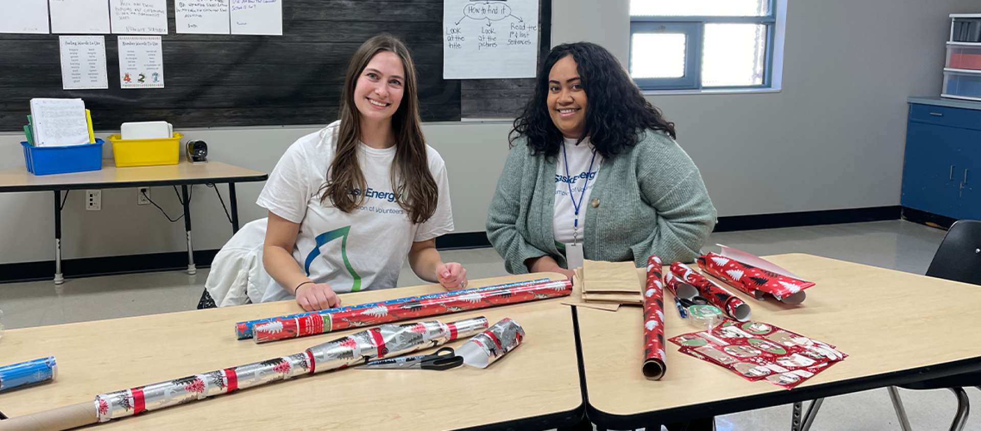 Two women with long hair smile, seated at a table with tubes of red wrapping paper in front of them.paper