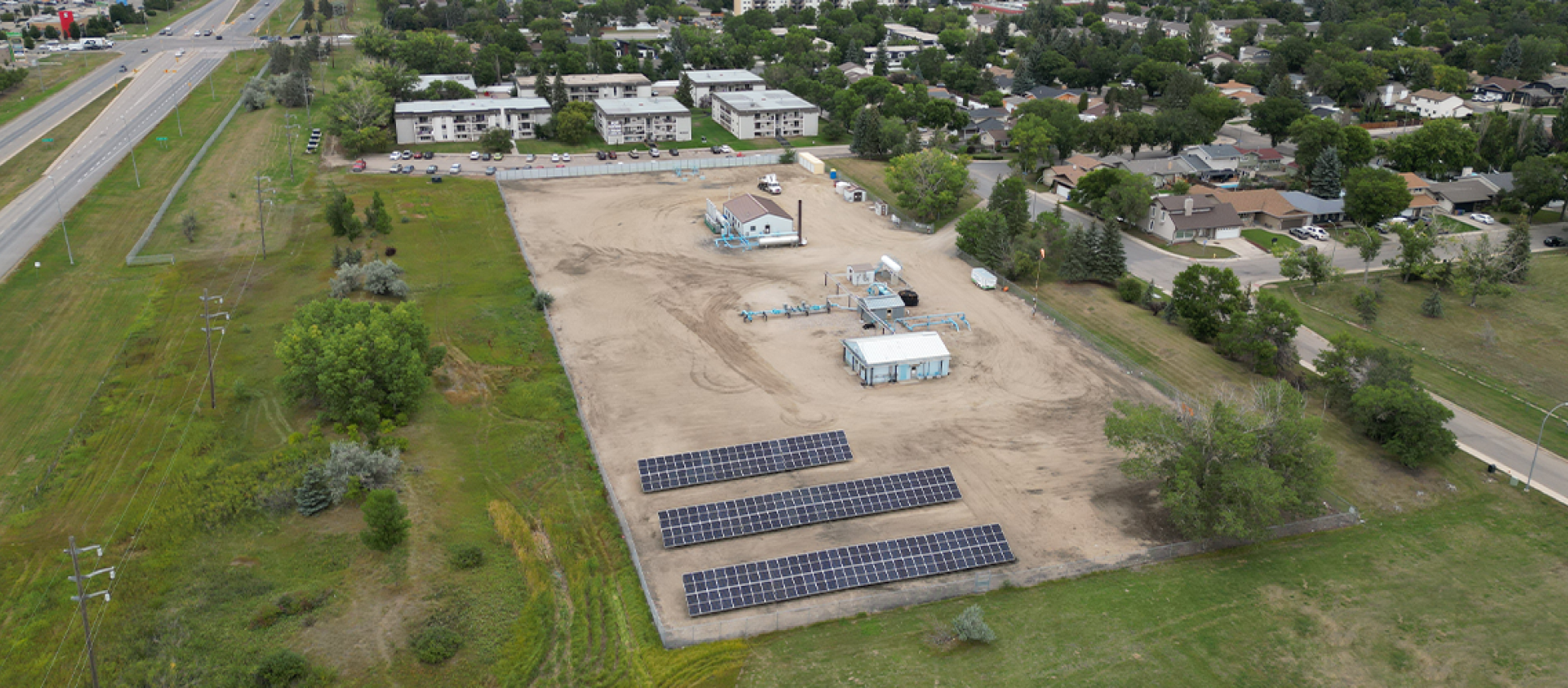aerial image - three rows of solar panels at centre, surrounded by light brown earth, grass and trees, houses and roads.