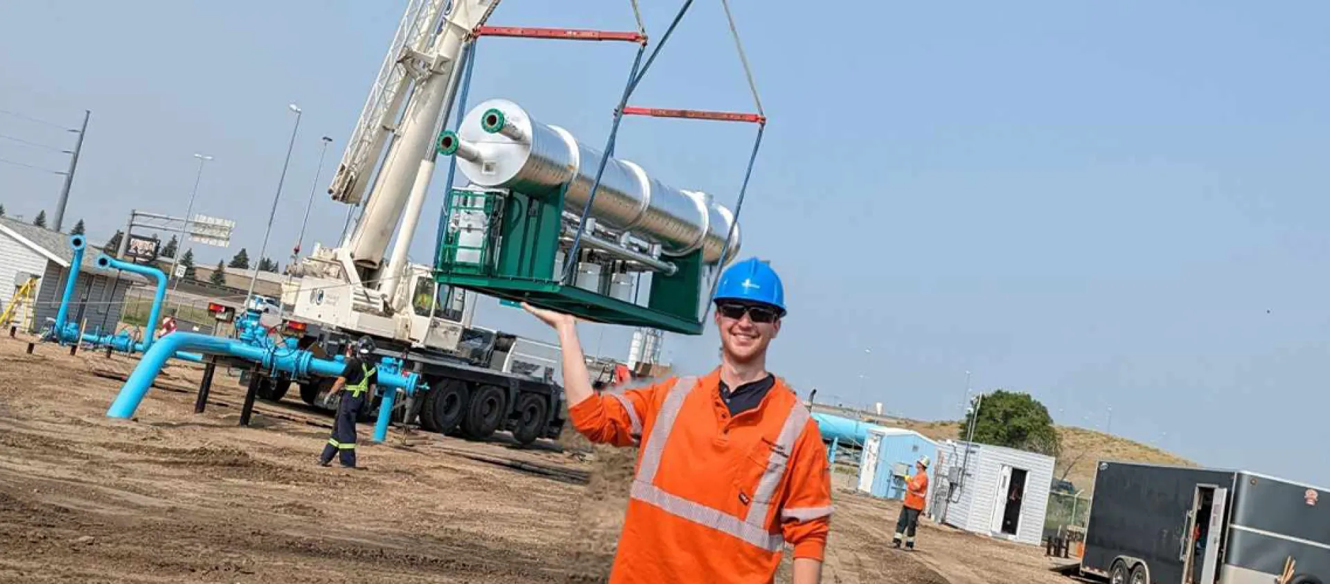 A man wearing an orange workshirt and blue hard hat holds up his arm, to make it appear he is lifting a large silver line heater on a crane in the background.