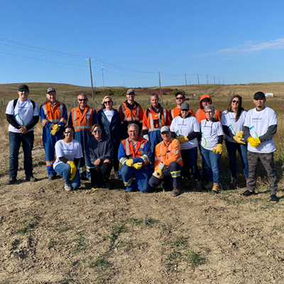 16 people - many wearing orange work clothes - stand or kneel in a posed group photo, with blue sky above and brown grass around them