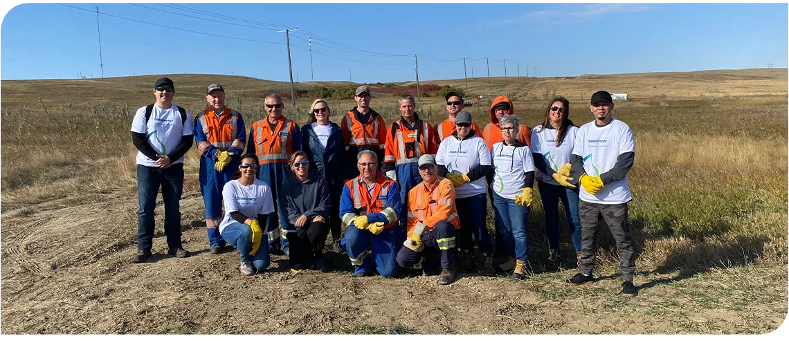 16 people - many wearing orange work clothes - stand or kneel in a posed group photo, with blue sky above and brown grass around them