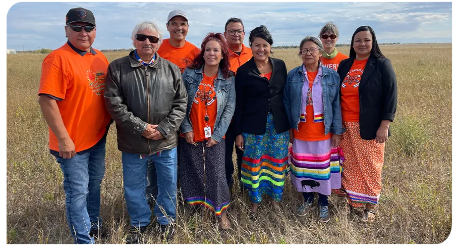 Nine people stand in a row outdoors in a golden field. Many wear orange T-shirts and/or colourful ribbon skirts.