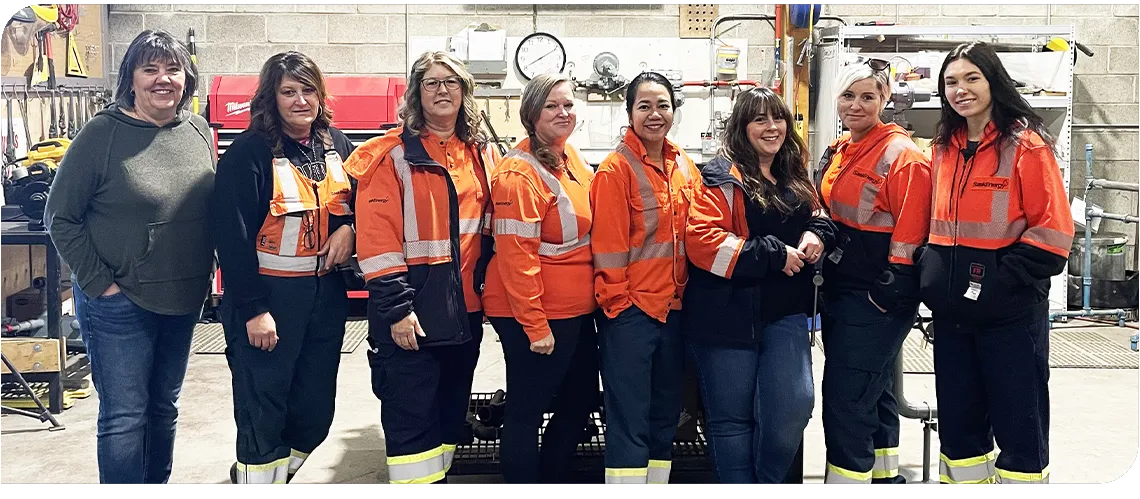 Eight women stand in a row in a shop with tools behind. Seven of them are wearing orange and navy work clothes. 
