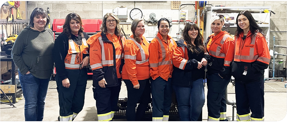 Eight women stand in a row in a shop with tools behind. Seven of them are wearing orange and navy work clothes.